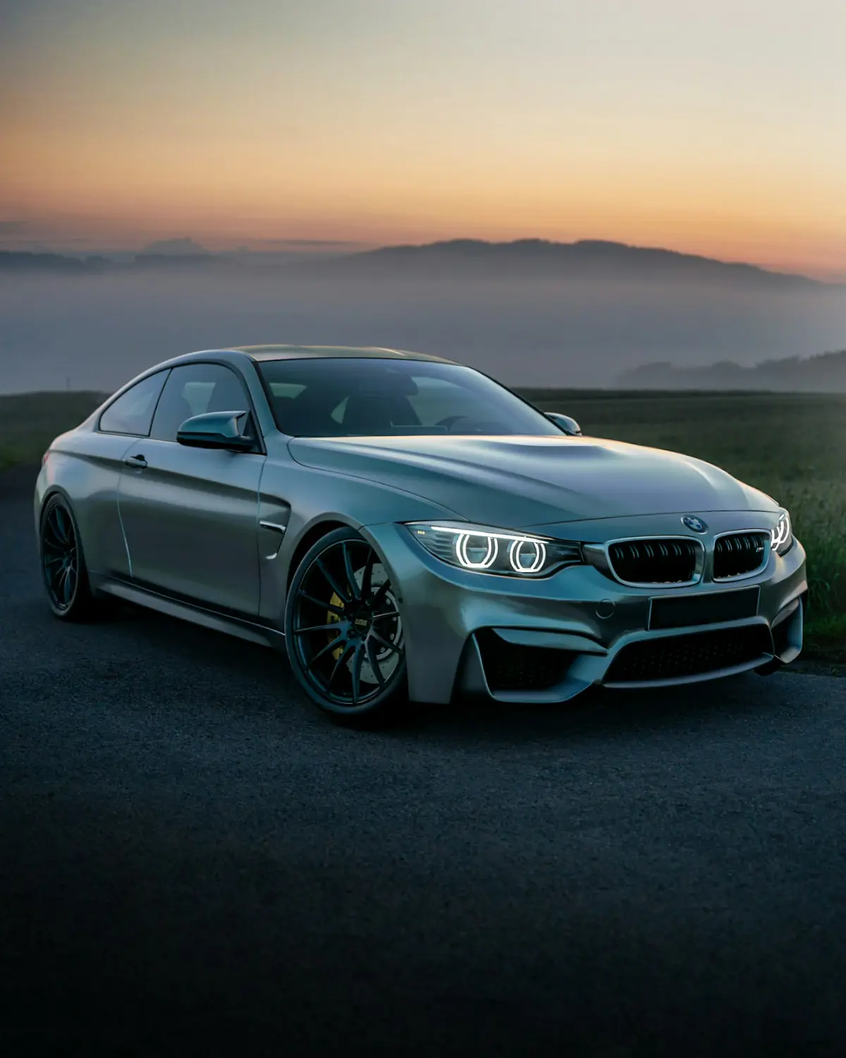 Grey BMW M4 at dawn with headlights on, misty hills and an amber horizon behind